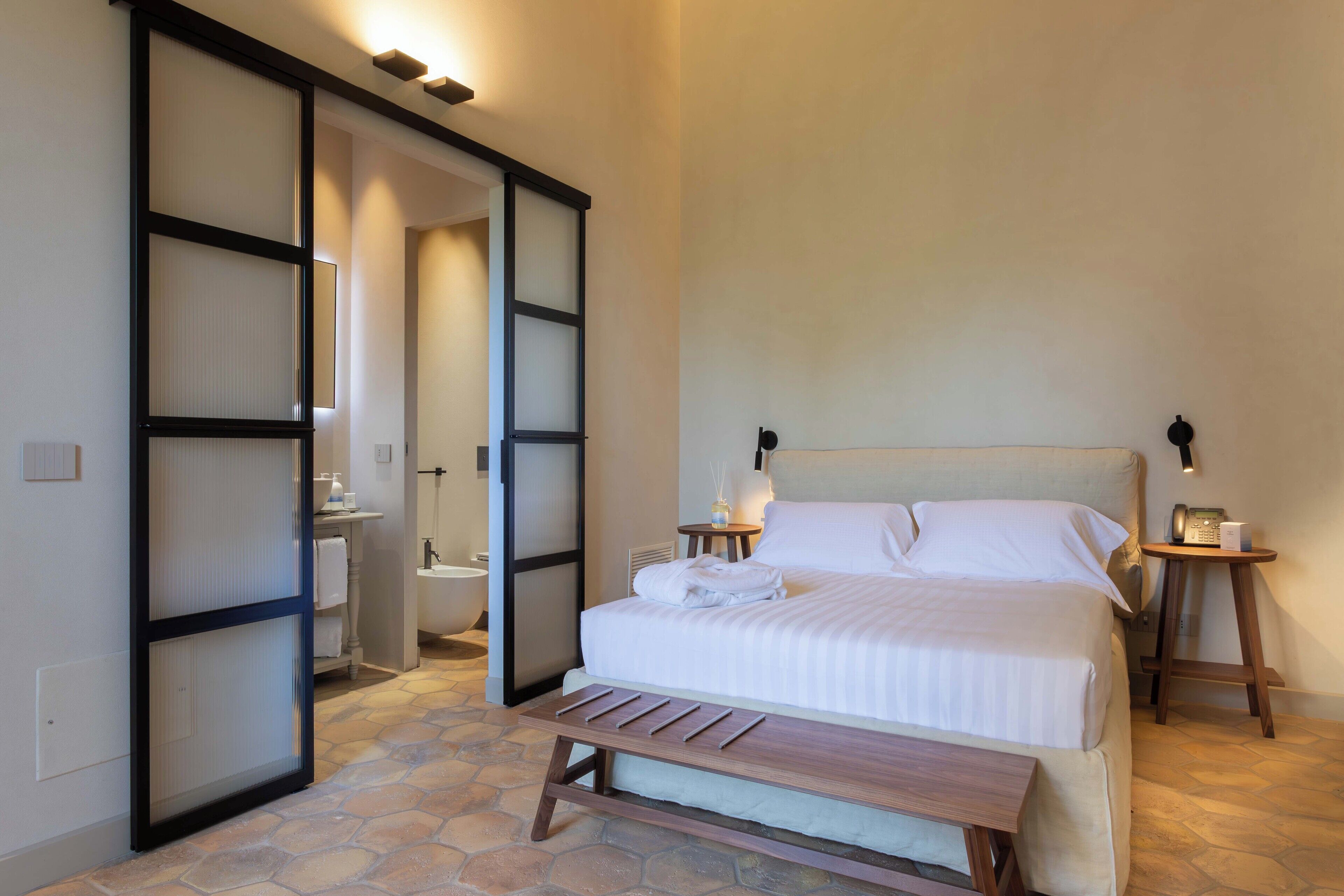 A bedroom with a bed, bedside tables, a bench, a bathroom with a bathtub, and a sliding door.