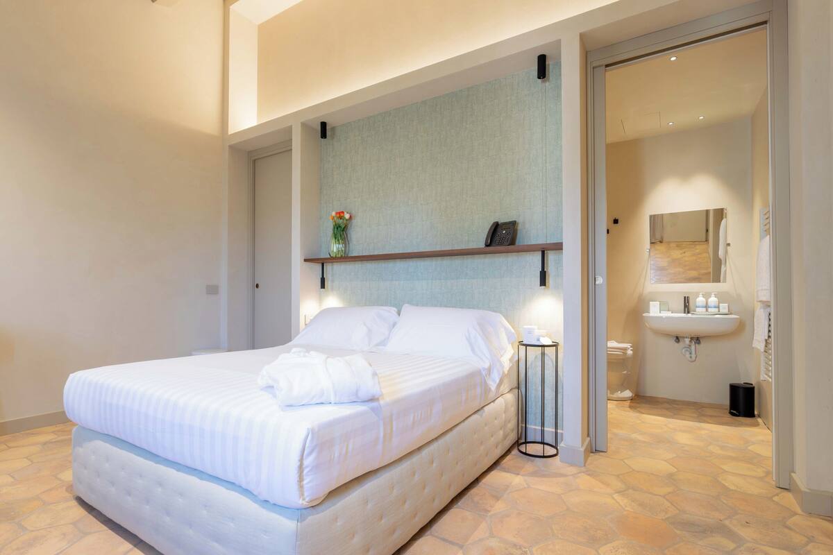 Un dormitorio con cama, mesita de noche y baño con lavamanos y espejo.