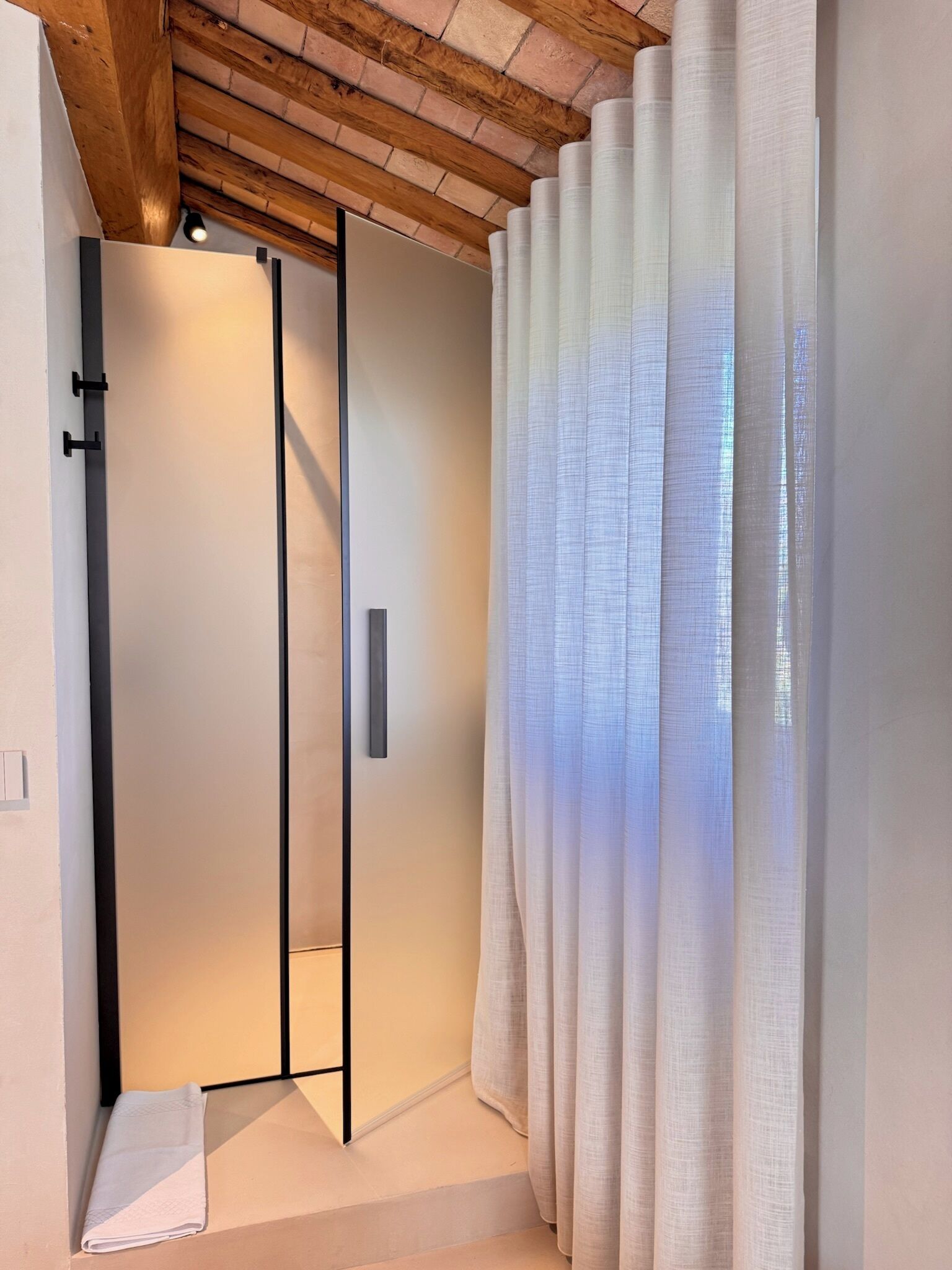 A modern bathroom with a glass shower enclosure, a towel on the floor, and a window with sheer curtains.