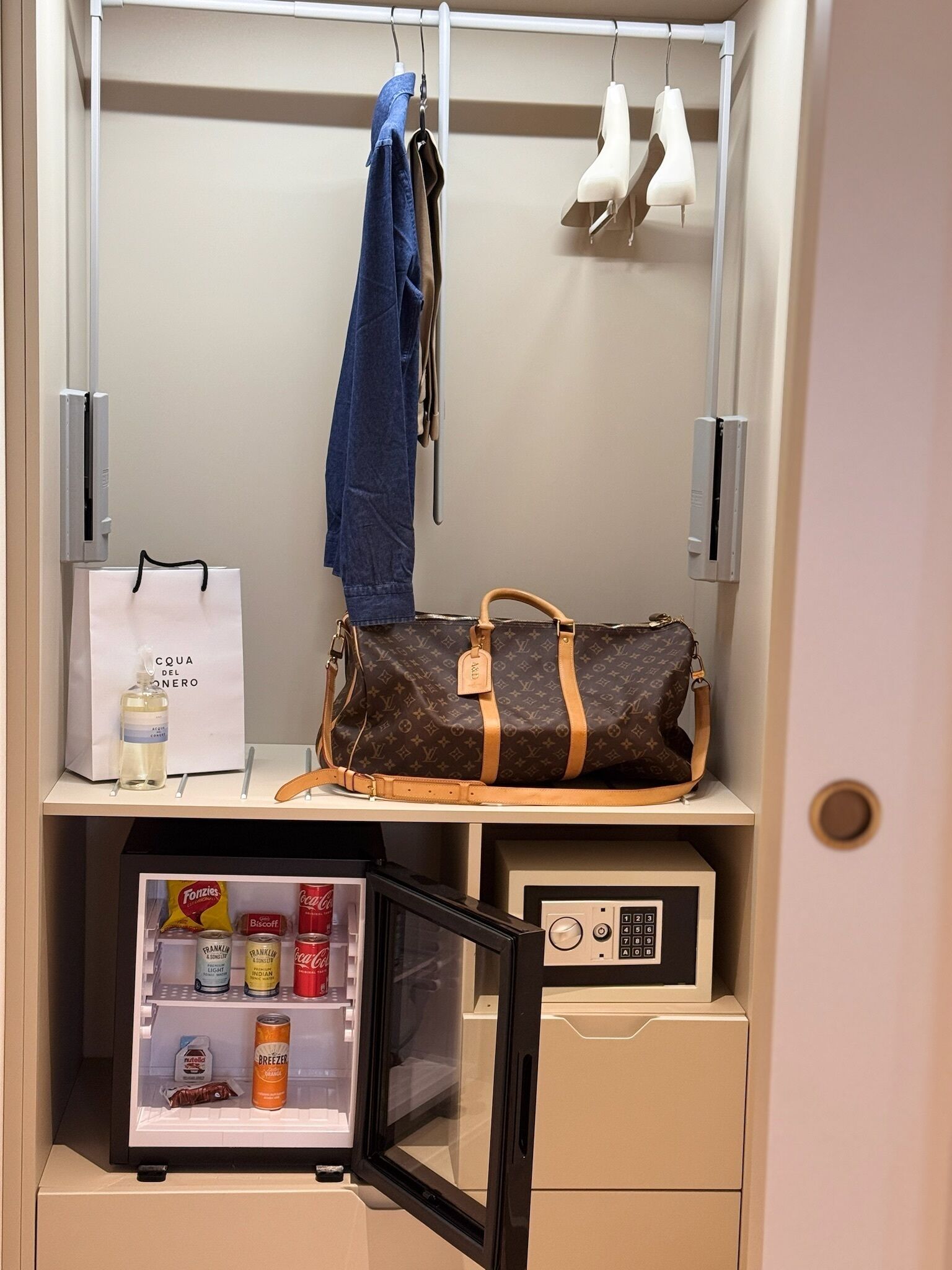 Een kast met hangende kleding, een handtas en een minibar gevuld met snacks en drankjes.