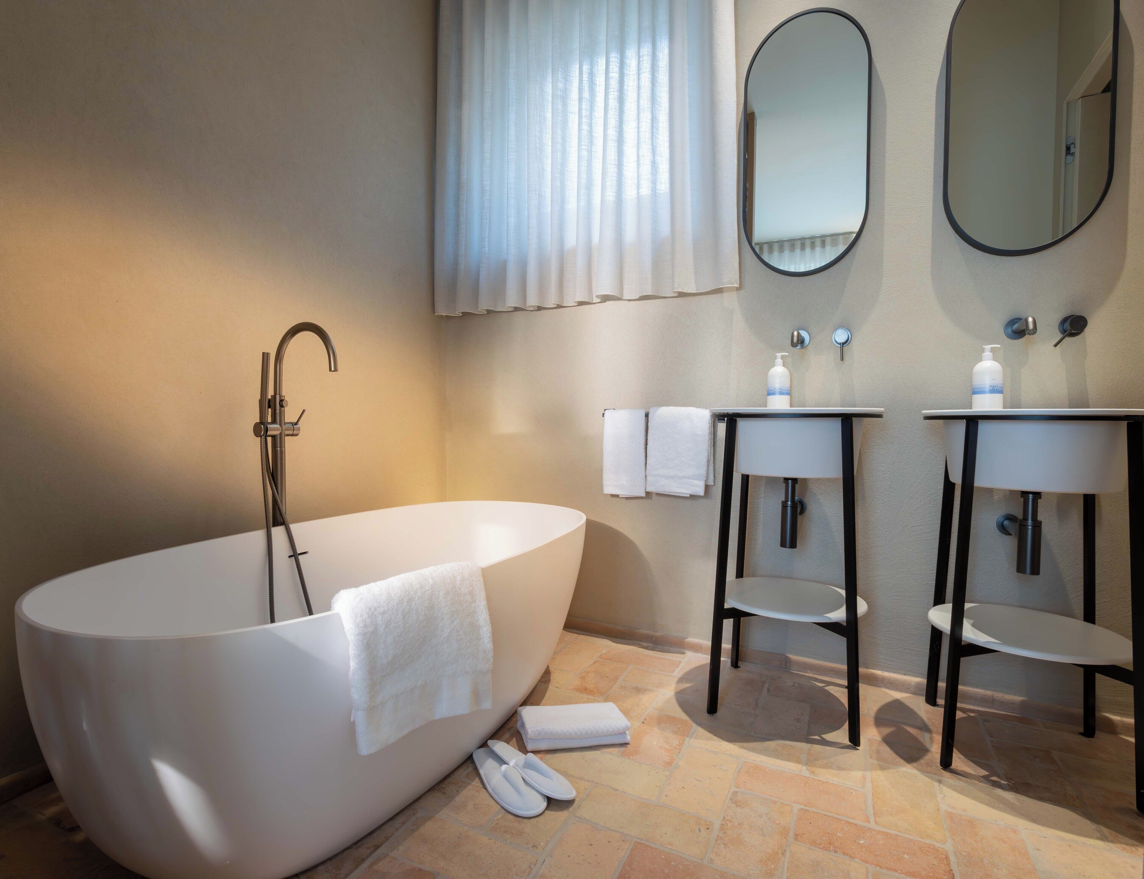 Un bagno moderno con una vasca freestanding, due lavandini, uno specchio e una finestra con tende.