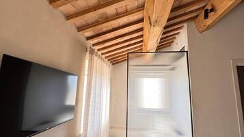 Una stanza con soffitto in legno, una televisione a schermo piatto appesa al muro, una finestra con persiane e un tavolino.