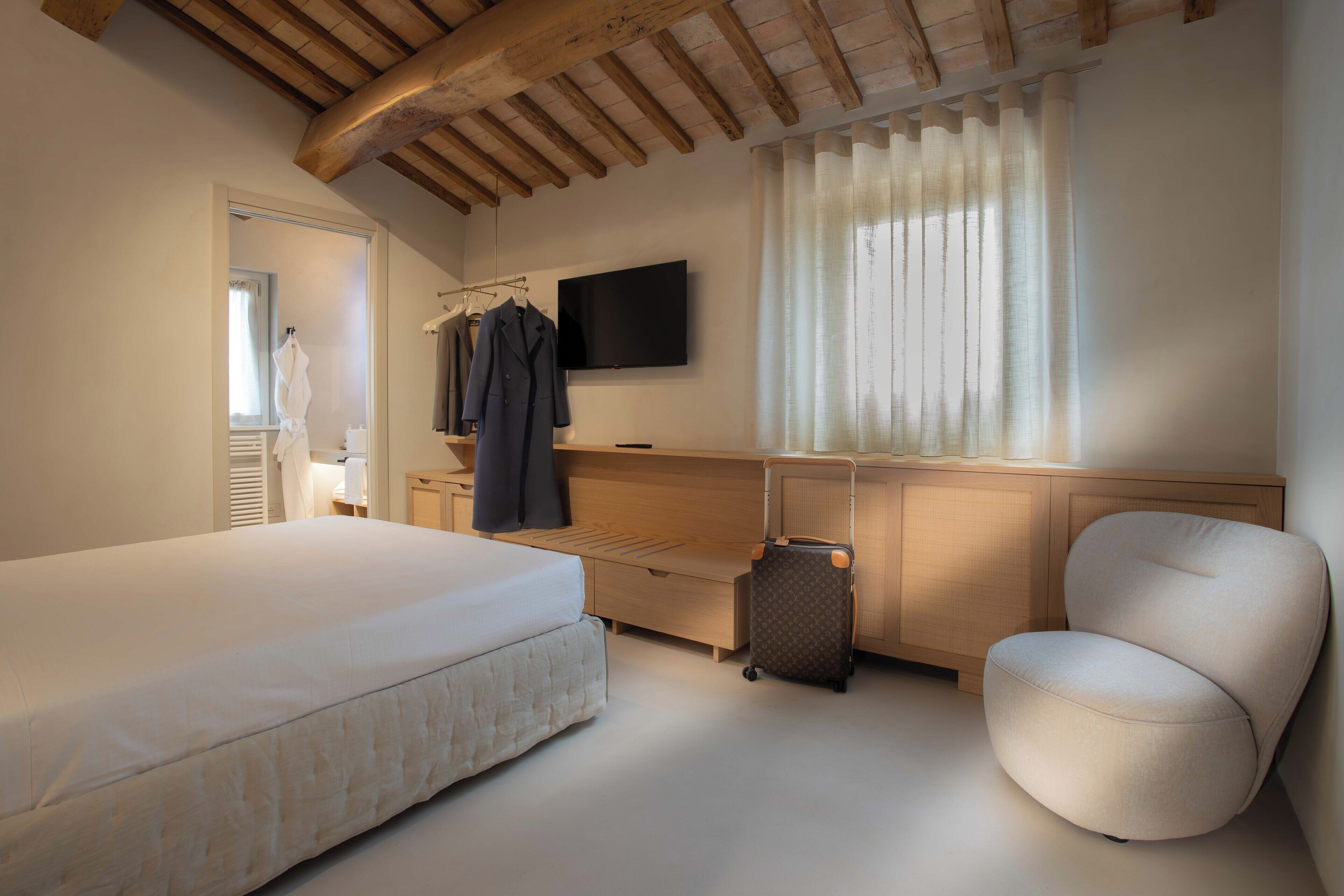 Una camera da letto con un letto, una sedia, una scrivania con televisore, una valigia e un appendiabiti.