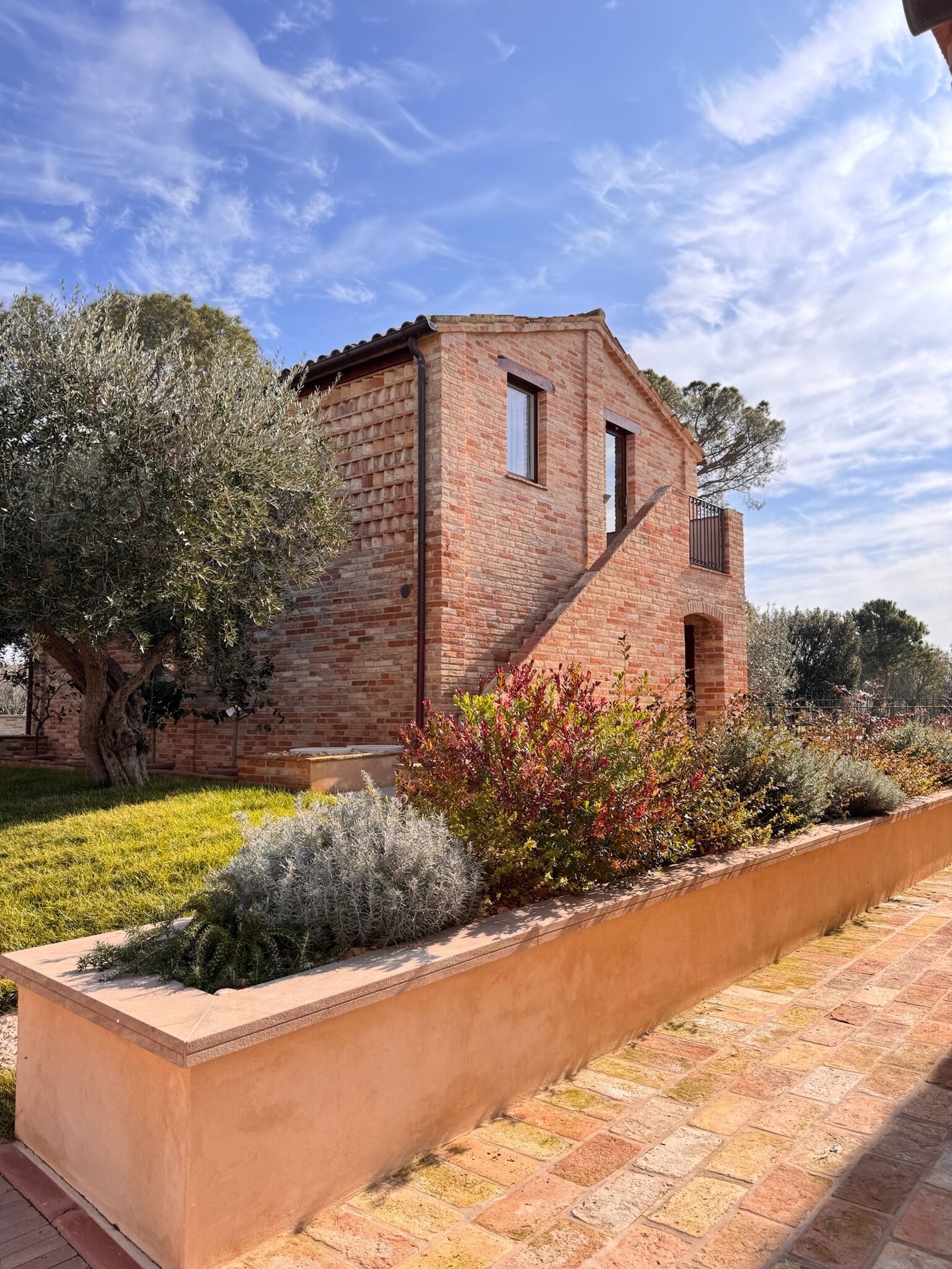 A brick house with a small balcony, surrounded by a garden with various plants and a tree.