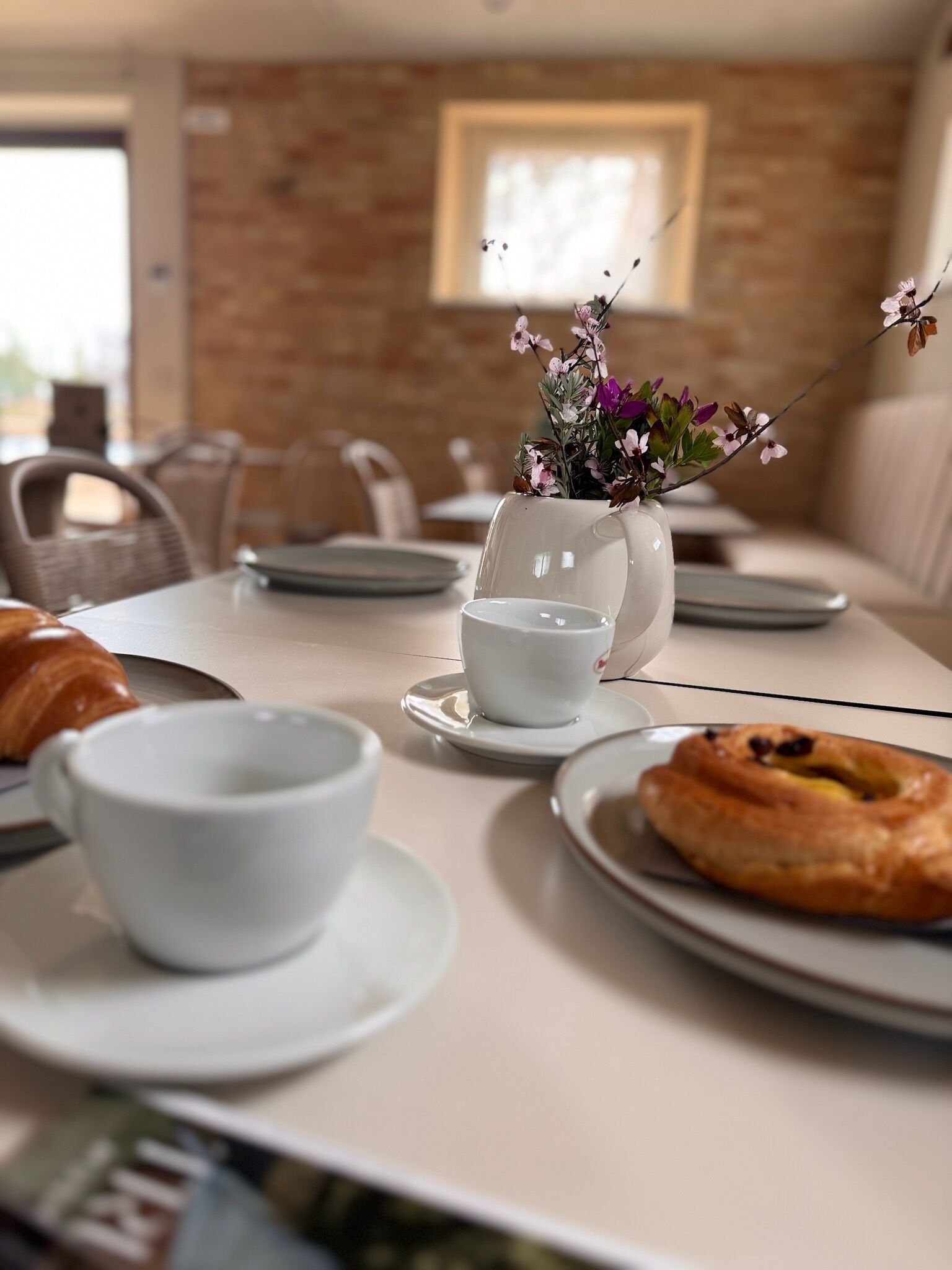 Aamiaispöydällä on maljakko täynnä kukkia, croissant ja lautanen ruokaa.