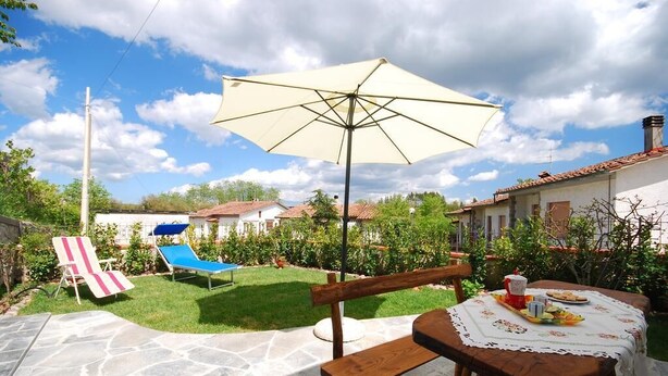 Un patio con una mesa de madera, una silla lounge azul, una silla a rayas y un paraguas grande.