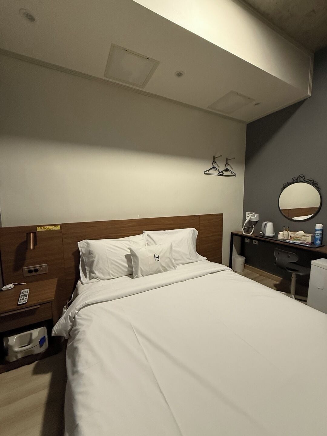 Habitación de hotel con una cama bien hecha, una mesita de noche con lámpara, un pequeño escritorio con espejo y un zapatero de pared.