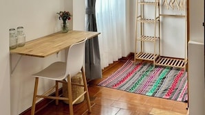 A small, bright room with a wooden desk, a white chair, a small table with a vase of flowers, a striped rug, and a metal drying rack. - La Maison Samaï (Urubamba)