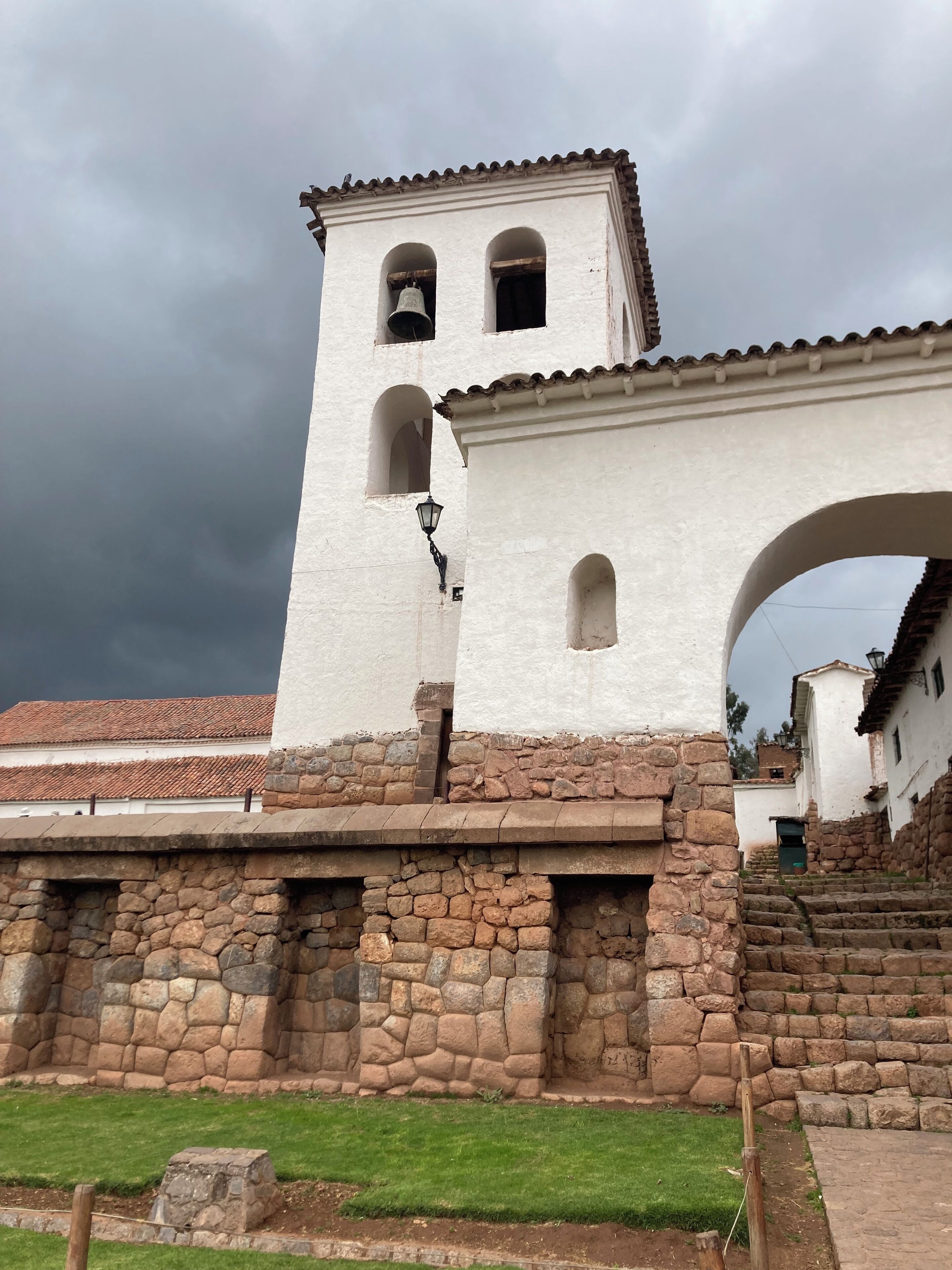 A white building with a bell tower and arched windows, surrounded by stone walls and a grassy area.