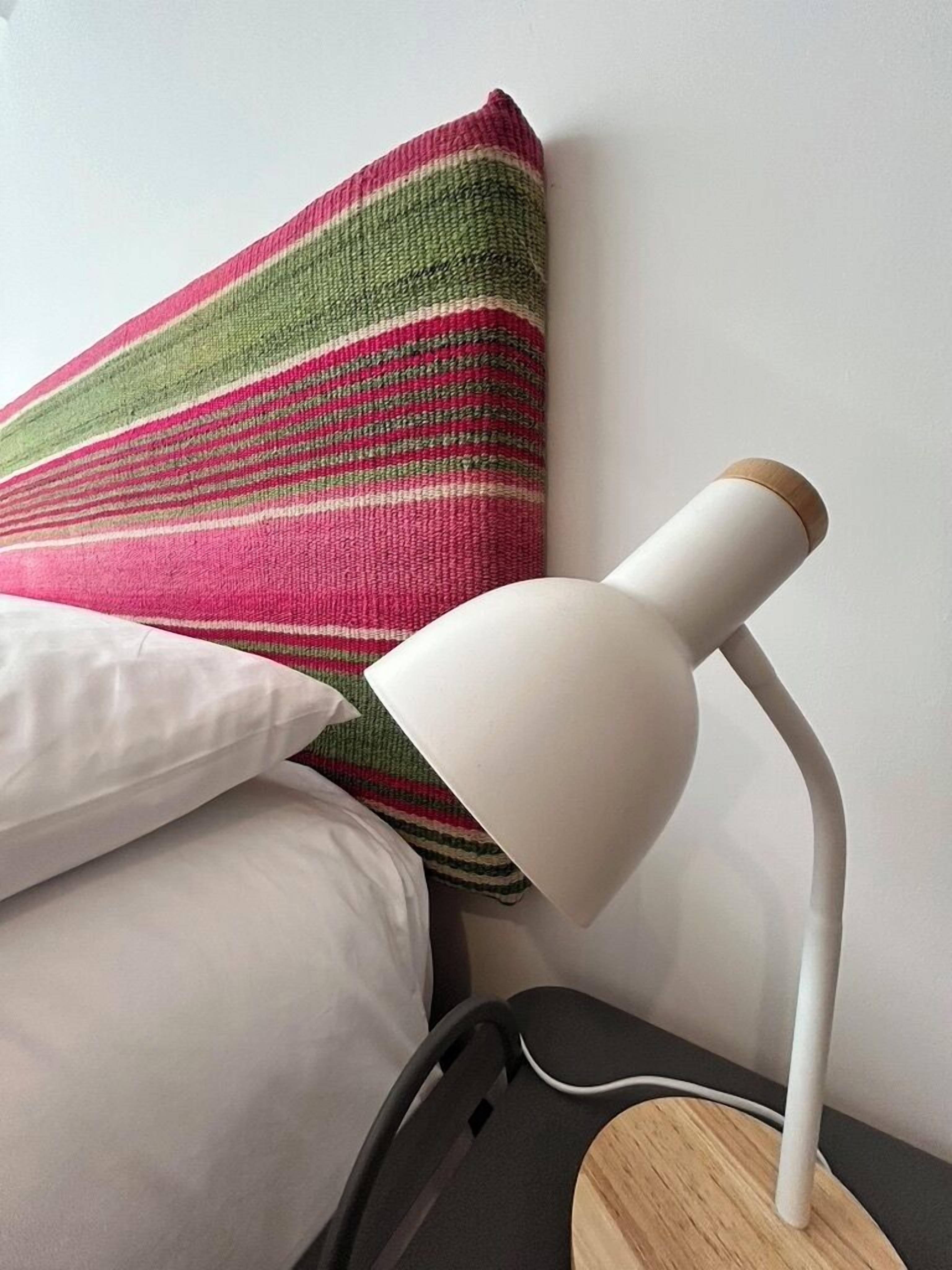 Una cama con almohadillas blancas y una manta a rayas de colores, una lámpara de escritorio blanca con base de madera y una silla negra.