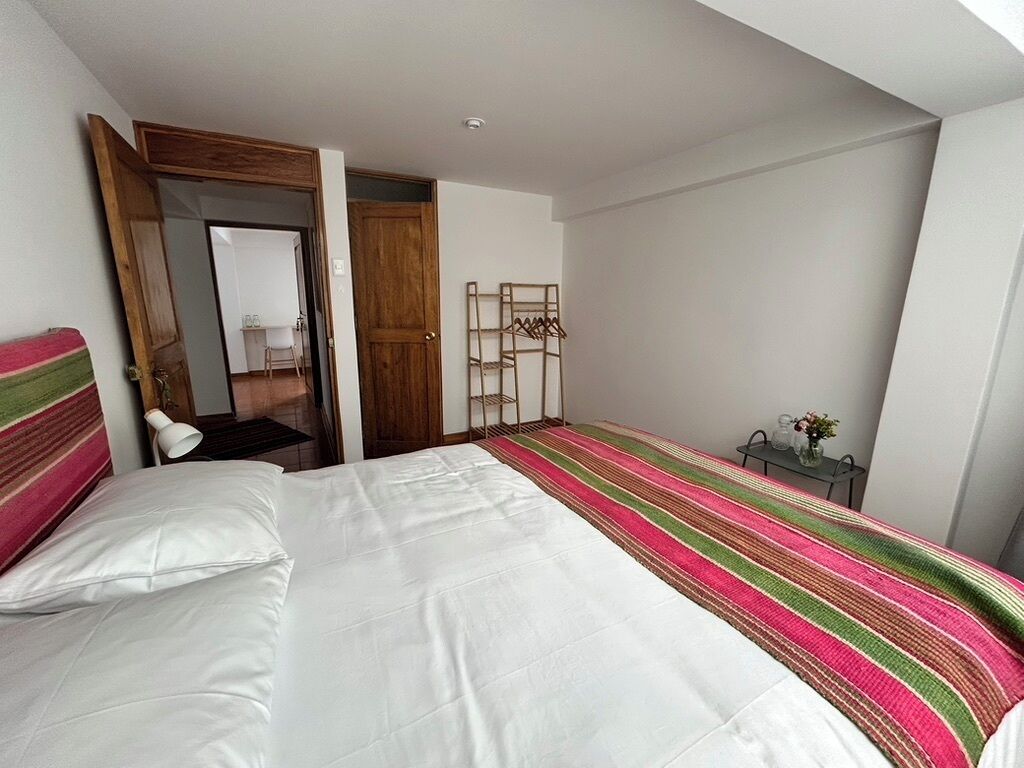 A bedroom with a bed, a wooden door, a coat rack, and a small table with a vase.