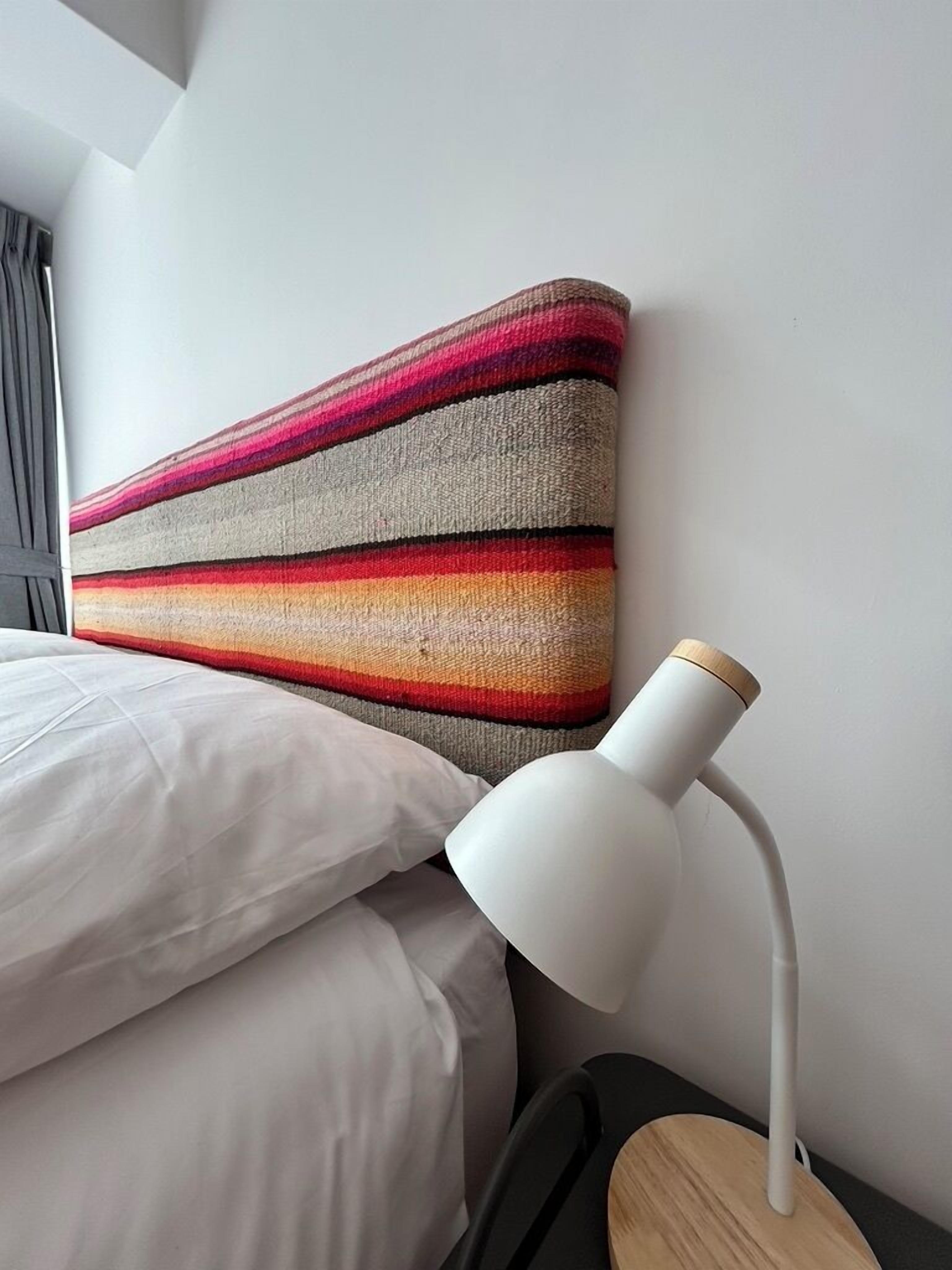 Una cama con sábanas blancas, un tapiz de pared a rayas de colores y una lámpara de escritorio blanca.