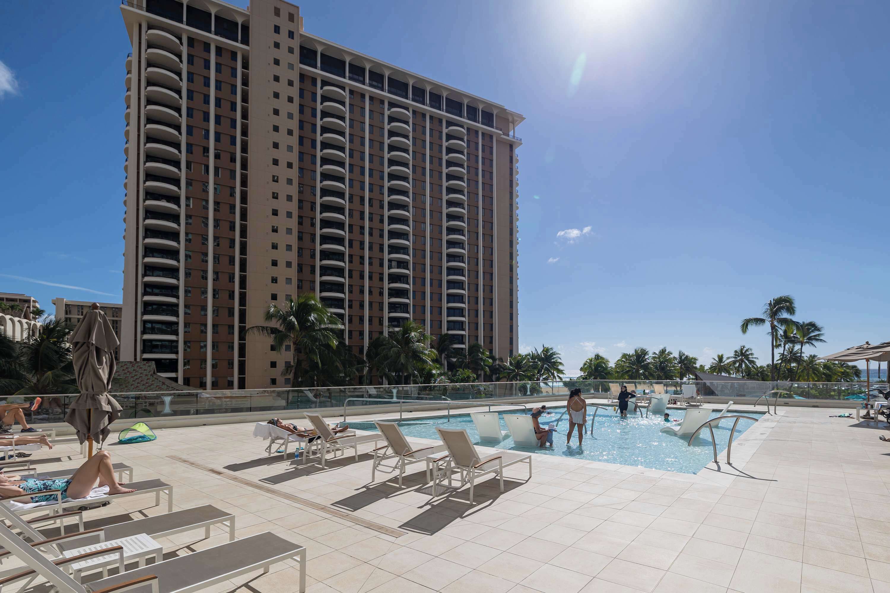 A hotel pool area with sun loungers, a large building in the background, and palm trees.