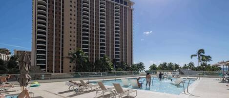 A hotel pool area with sun loungers, a large building in the background, and palm trees.