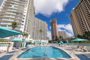 A swimming pool area with lounge chairs and umbrellas, surrounded by tall buildings.