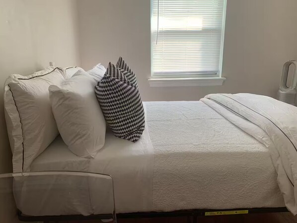 A single bed with white bedding and a patterned pillow, a bedside table, and a window with blinds.