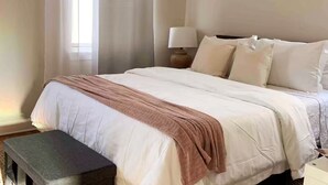 Una cama bien hecha con una manta blanca y marrón, una lámpara de noche y un pequeño banco.