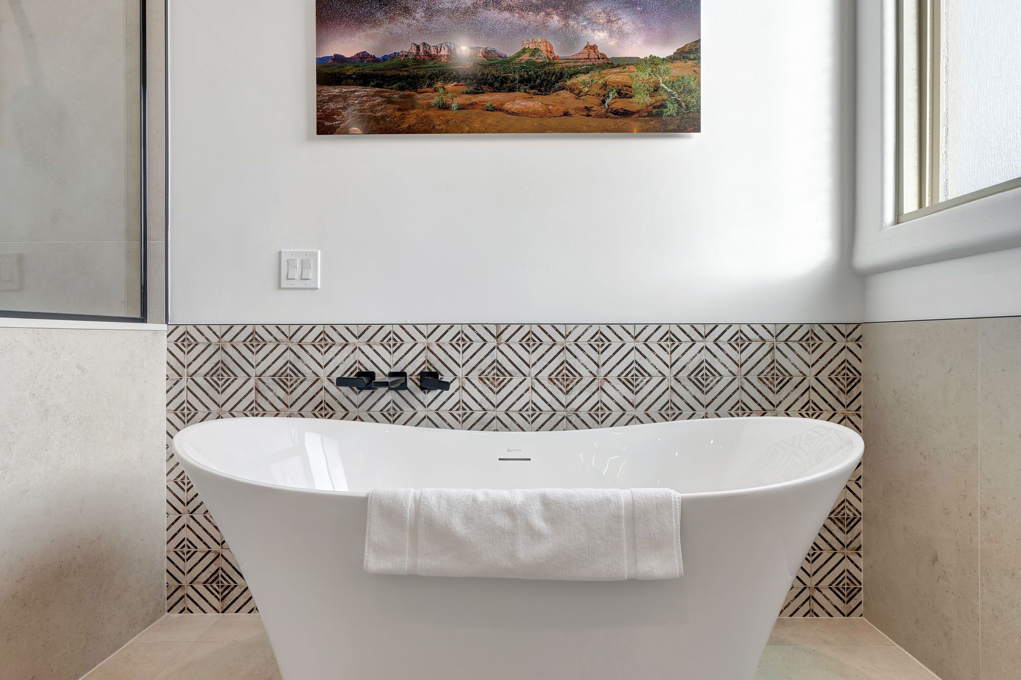 A bathroom with a freestanding bathtub, patterned wall tiles, and a framed landscape painting above the tub.