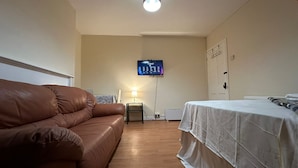 Une pièce avec un canapé en cuir marron, un lit blanc, un téléviseur à écran plat fixé au mur, une lampe sur une petite table et un luminaire au plafond.