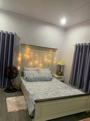A bedroom with a bed, curtains, a nightstand, a lamp, and a fan. - Ruby House, 3 Bedroom Apartment, Beachside (La Gi)