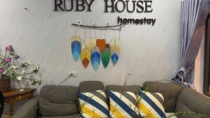 A living room with a sofa, decorative pillows, a wall-mounted sign reading "RUBY HOUSE homestay", and a branch with leaves. - Ruby House, 3 Bedroom Apartment, Beachside (La Gi)