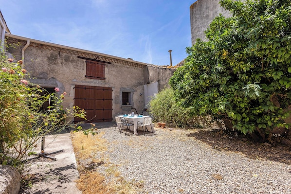 Un bâtiment rustique avec une cour en gravier, une table entourée de chaises et une grande porte en bois.