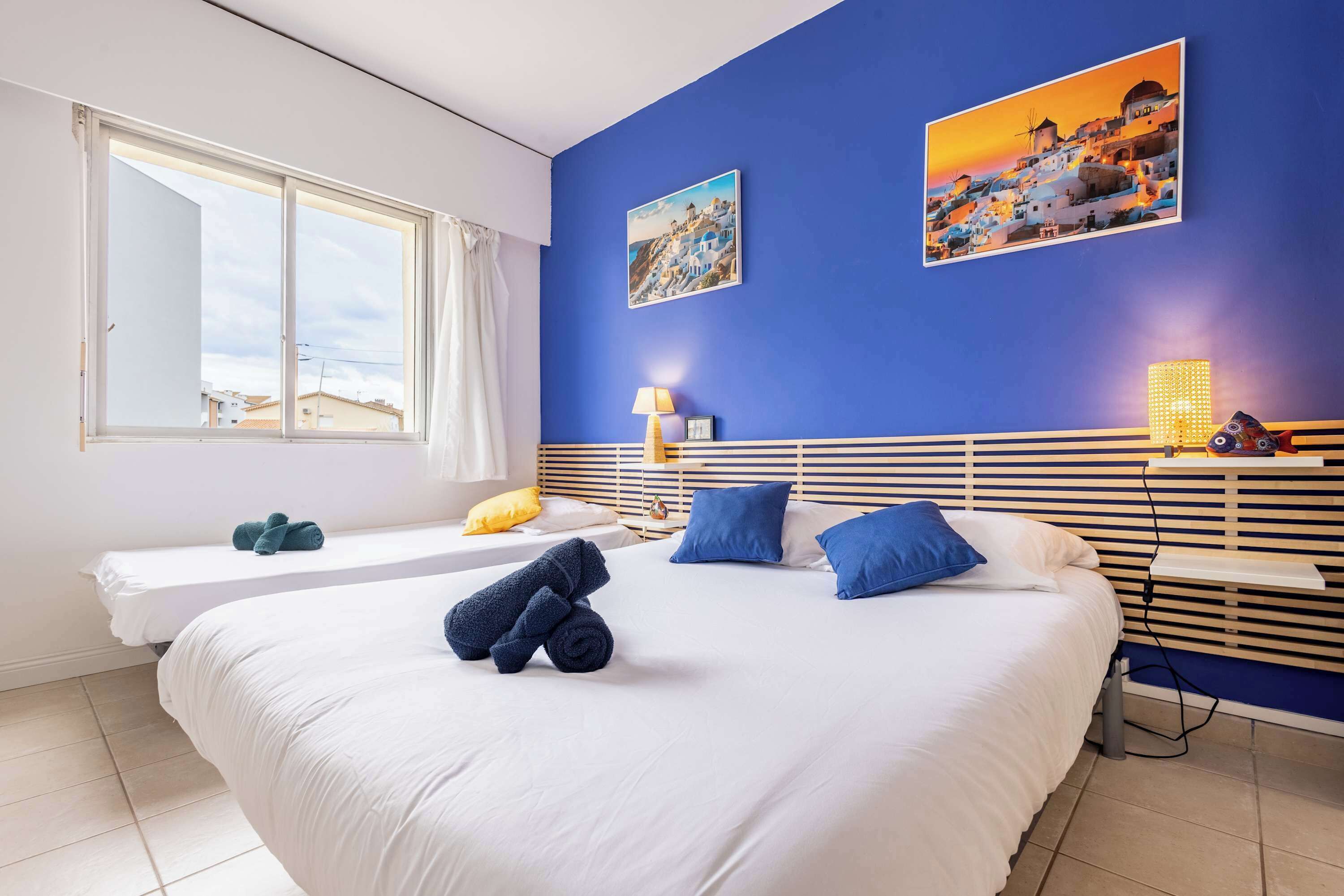 Een hotelkamer met een groot bed, een kleiner bed, een raam met gordijnen, een wandlamp en twee schilderijen.