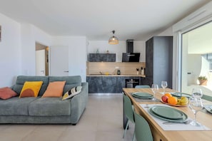 A modern living room with a grey sofa, a dining area with a wooden table set for two, and a kitchen in the background. - Valras'Loc Archipel Panarea Horizon Bleu (Valras-Plage)