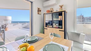 A dining area with a table set for two, a cabinet with dishes, and a view of the sea.