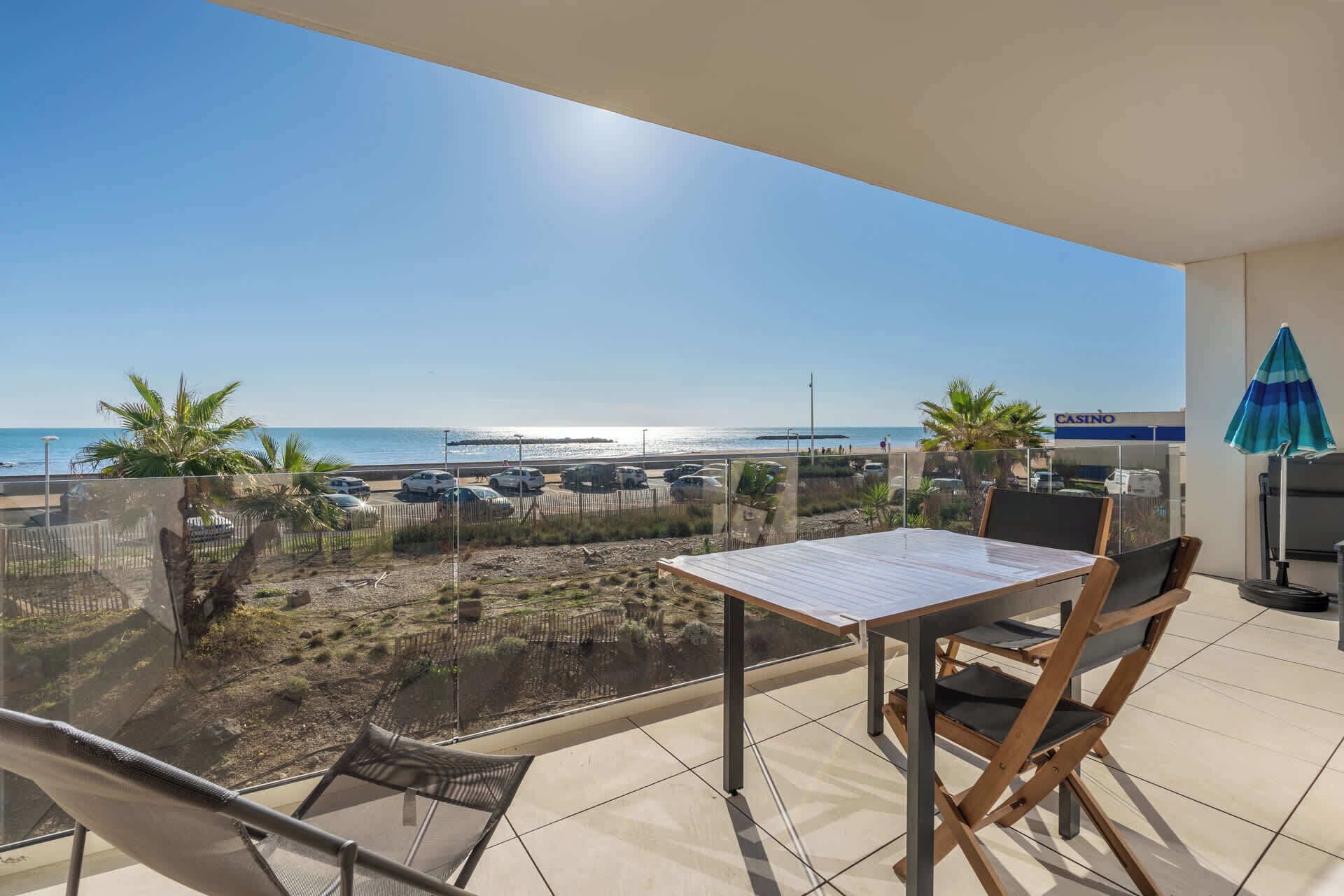 Een balkon met een tafel en stoelen, uitkijkend op een strand met geparkeerde auto's en een bord van een casino.