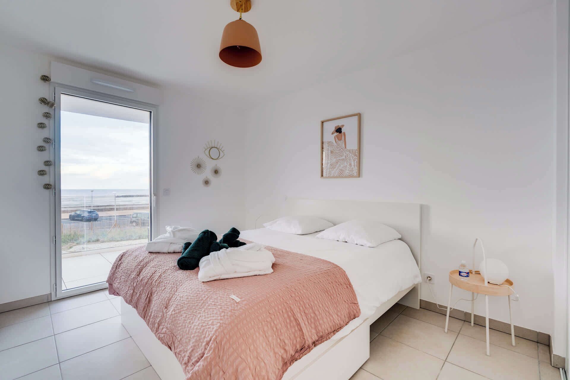 A modern bedroom with a large bed, a small bedside table, a hanging light fixture, and a view of the beach through the window.