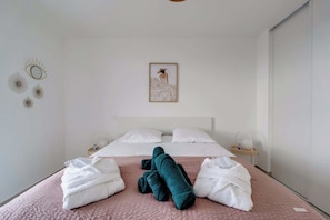 Habitación de hotel con una cama, dos almohadas blancas, una colcha rosa y dos toallas (una blanca y otra verde azulada).