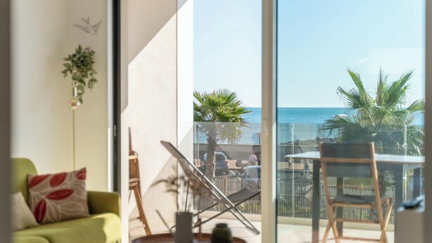 A room with a green sofa, a small table with a vase, and a chair. There is a large sliding glass door leading to a balcony with a view of the sea and palm trees. - Valras'Loc Archipel Capri Balcony over the Sea (Valras-Plage)