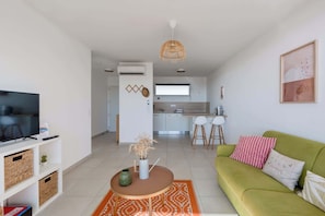 Una sala moderna con un sofá verde, una mesa de centro y un televisor. Se observa una zona de cocina al fondo con una mesa de comedor y sillas.