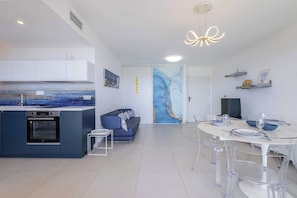 A modern kitchen with a built-in oven, a dining area with a round table and clear chairs, and a living space with a sofa and artwork.
