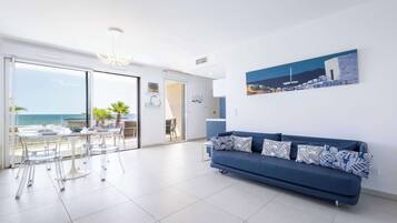 Een moderne woonkamer met een blauwe bank, een eettafel met stoelen en uitzicht op de zee door grote glazen deuren.