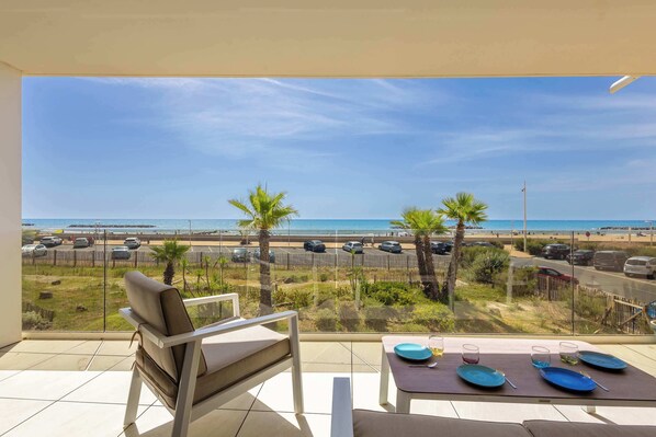 A balcony with a view of the beach, a table set for two, and a chair.