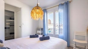 A bedroom with a bed, a hanging light fixture, a chair, and a view through a window.