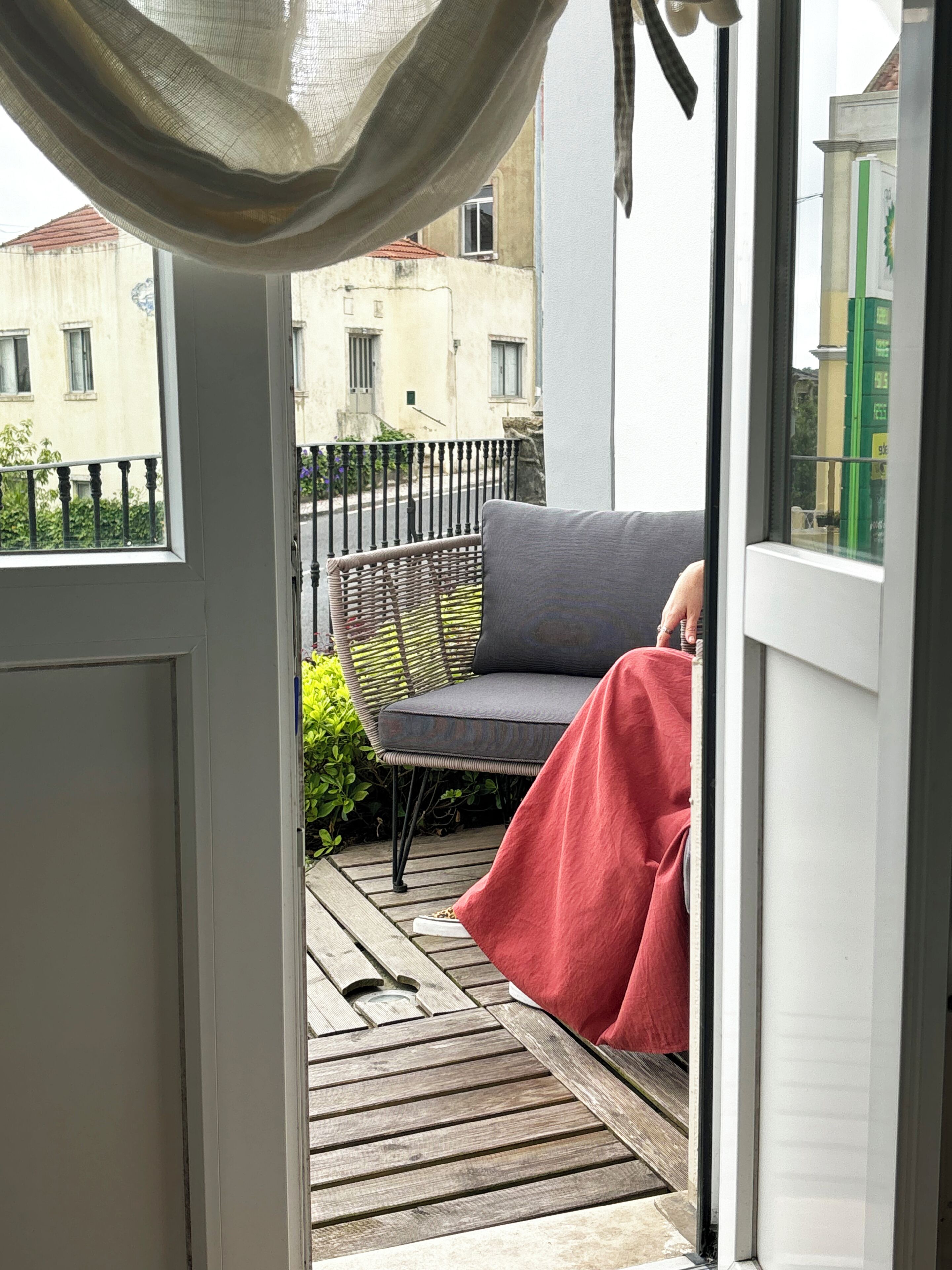 A balcony with a wicker chair and a red blanket, a wooden deck, and a view of buildings in the background.