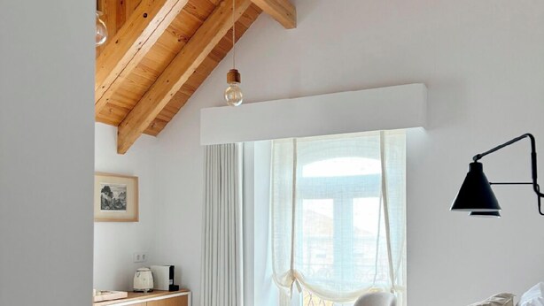 A bedroom with a wooden ceiling, a bed with a patterned bedspread, a bedside table with a lamp, and a window with sheer curtains. - Live Sintra Boutique Guest House (Sintra)
