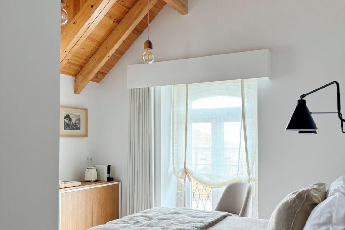 A bedroom with a wooden ceiling, a bed with a patterned bedspread, a bedside table with a lamp, and a window with sheer curtains.