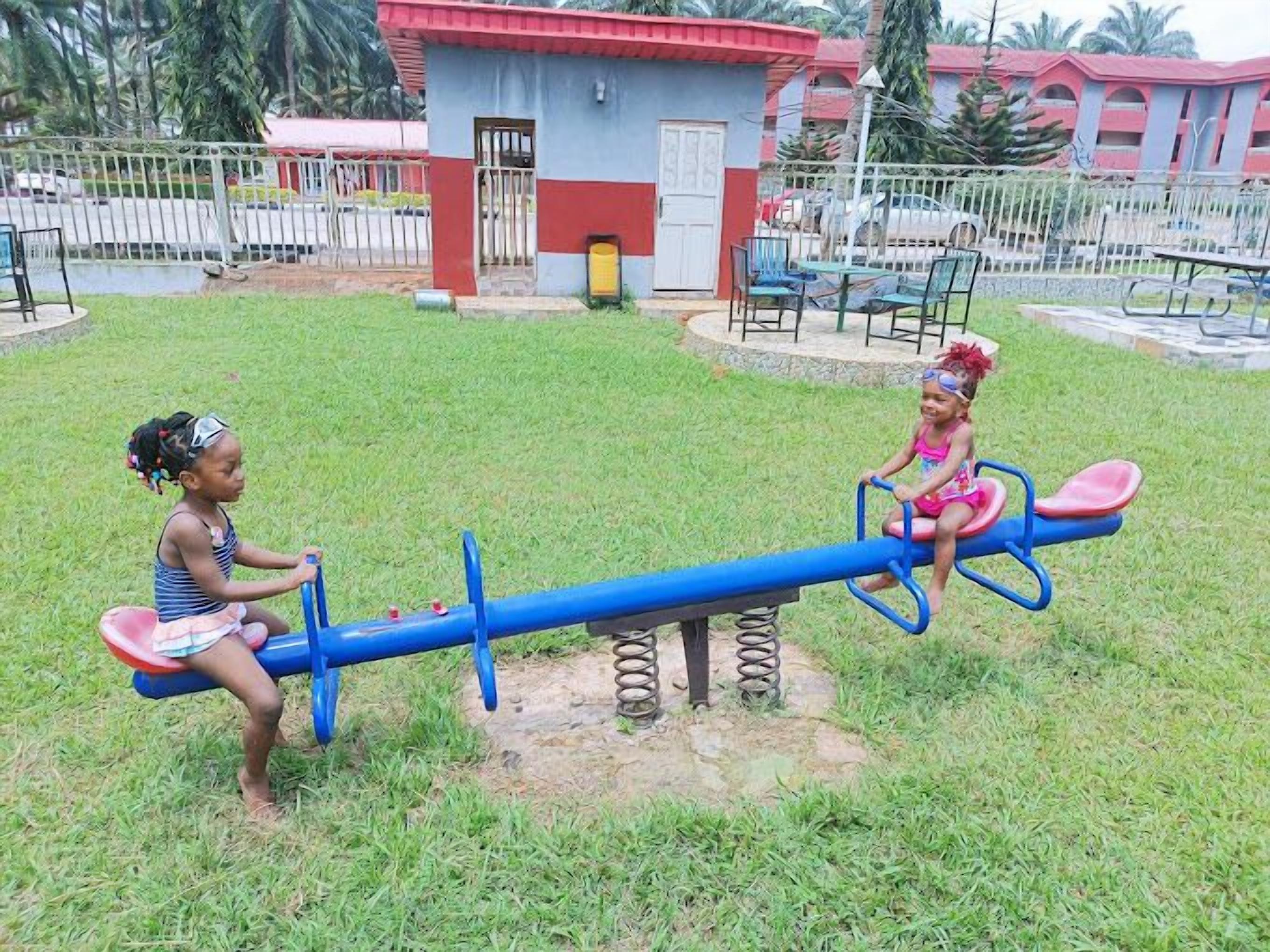 Deux enfants jouent sur une balançoire dans un parc. On aper çoit en arrière-plan un petit bâtiment et une zone délimitée par une clôture.