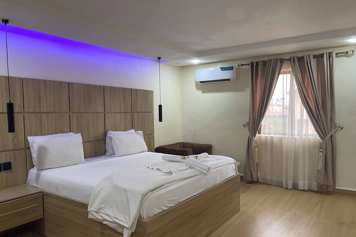 Ein modernes Schlafzimmer mit Holz-Kopfteil, einem Bett mit weißen Bettwäsche, einem Nachttisch, einer Klimaanlage und einem Fenster mit Vorhängen.
