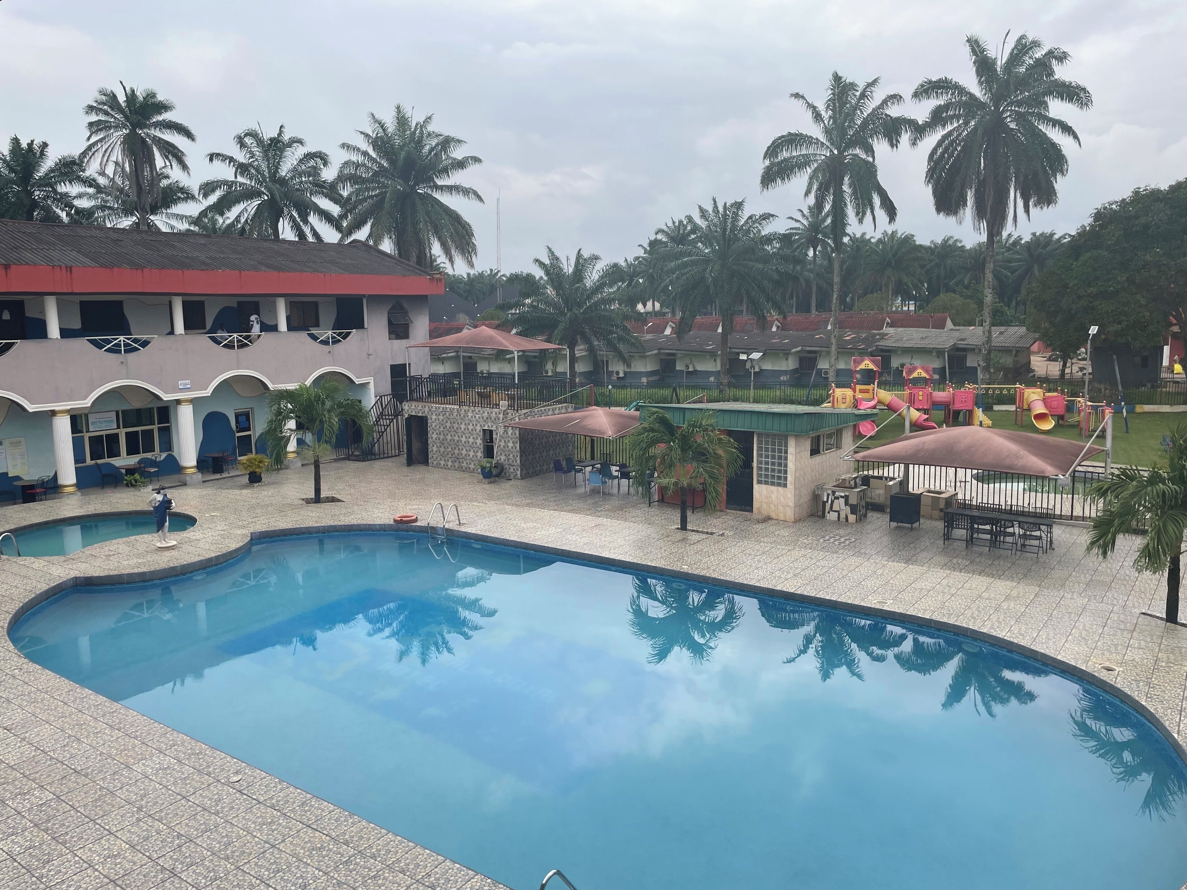 A swimming pool with a tiled deck, surrounded by palm trees and a playground with slides and play structures.