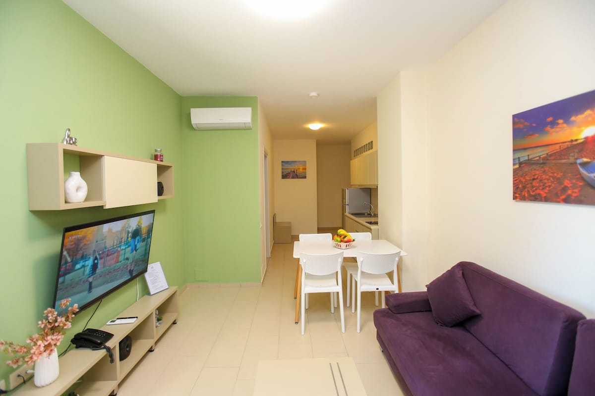 Un salon avec un téléviseur, un canapé violet, une table à manger avec des chaises, et un coin cuisine à l’arrière-plan.