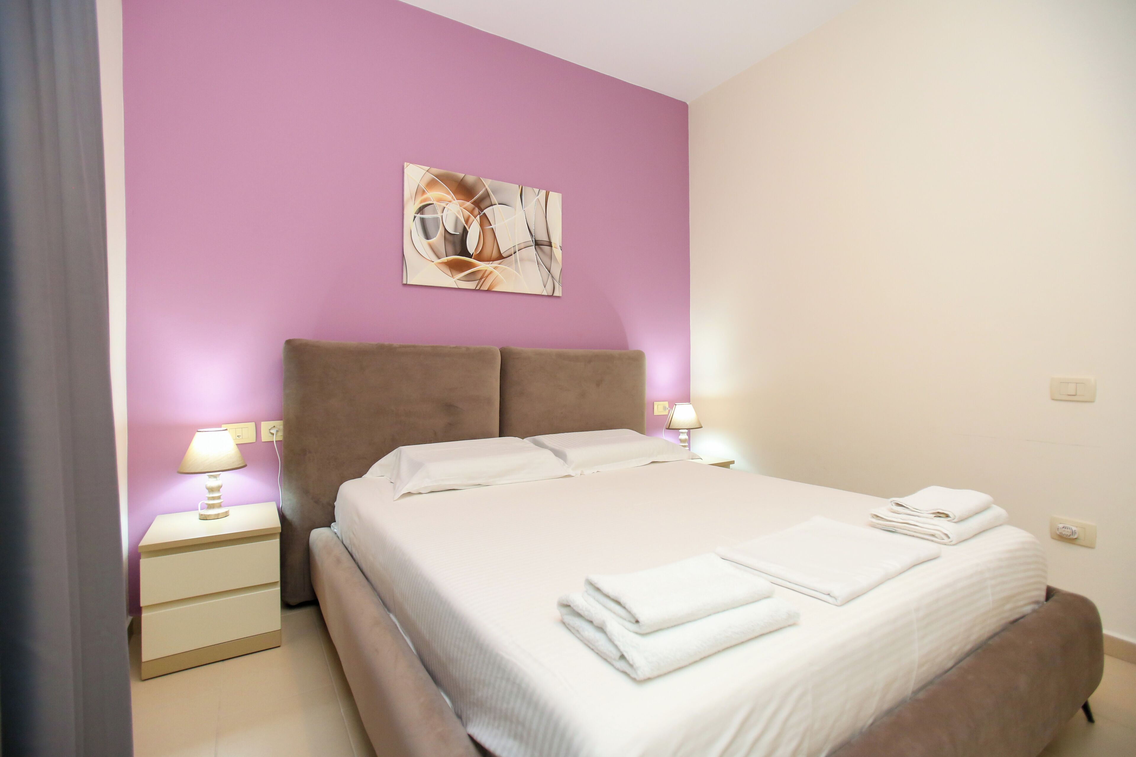 A neatly made bed with white linens and towels, a brown headboard, and a wall-mounted abstract painting.