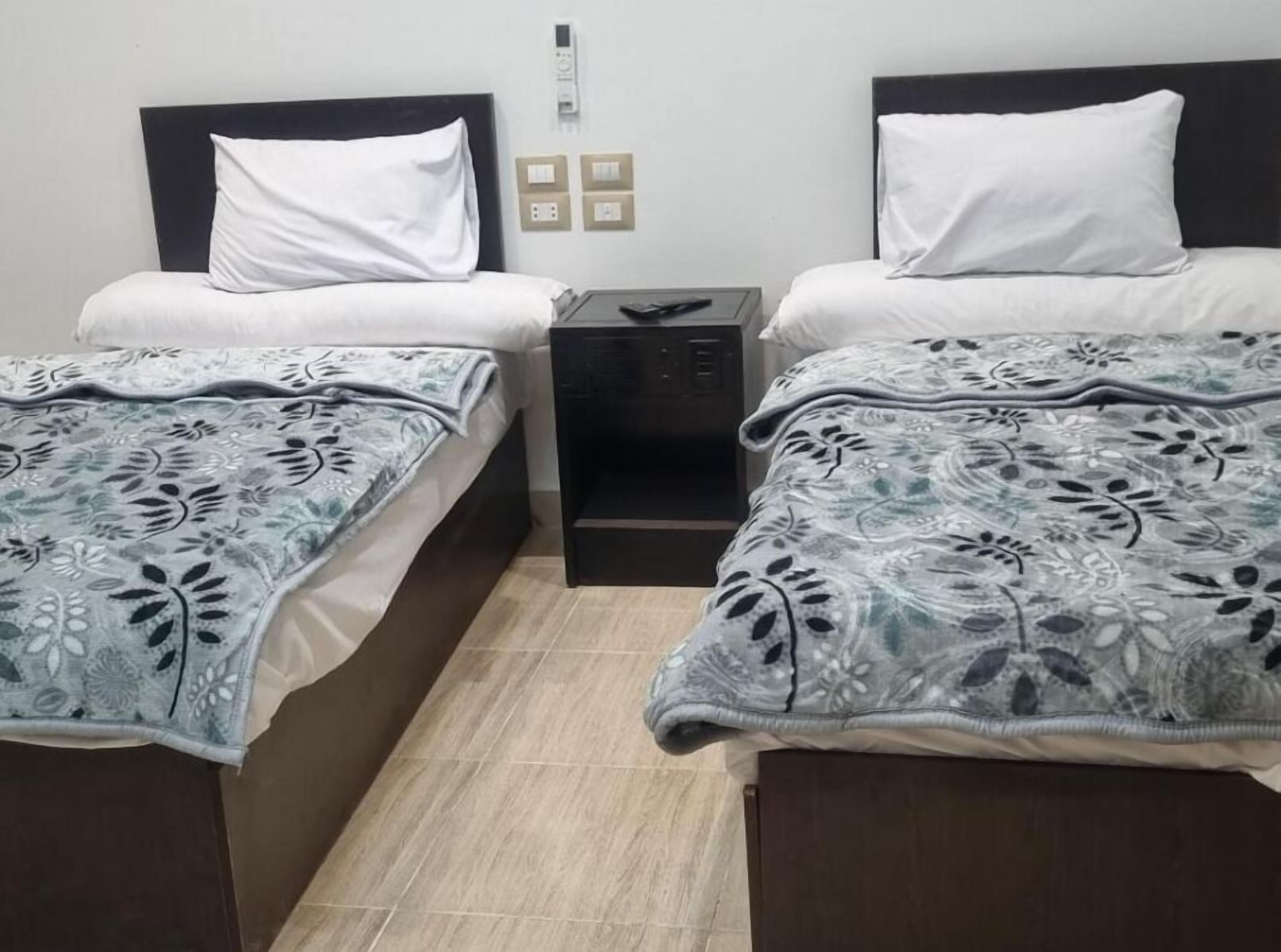 A twin room with two beds, each with a white pillow and a patterned blanket. There is a small bedside table with a lamp and a power outlet on the wall.
