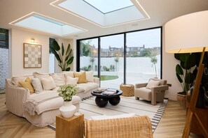 A modern living room with a sofa, armchairs, a coffee table, and a large window offering a view of the outside. - The Kensal Rise Retreat - Stunning 4bdr Home With Garden (London)