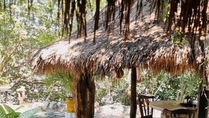 Un espacio de estar al aire libre cubierto, con techo de paja, bancos de madera y una pequeña mesa.
