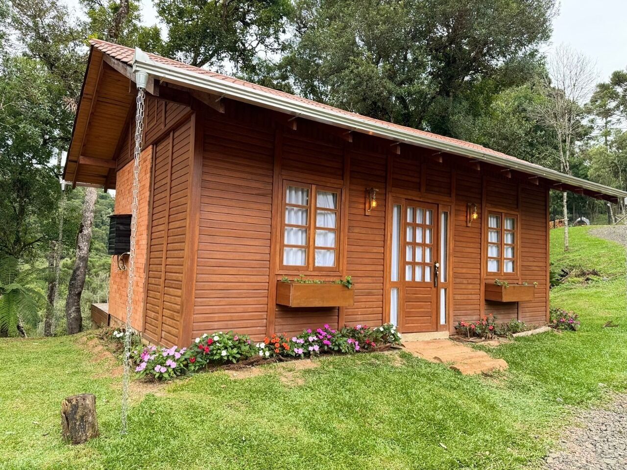 A wooden cabin with a sloped roof, two windows with curtains, and a small porch area with flower beds.